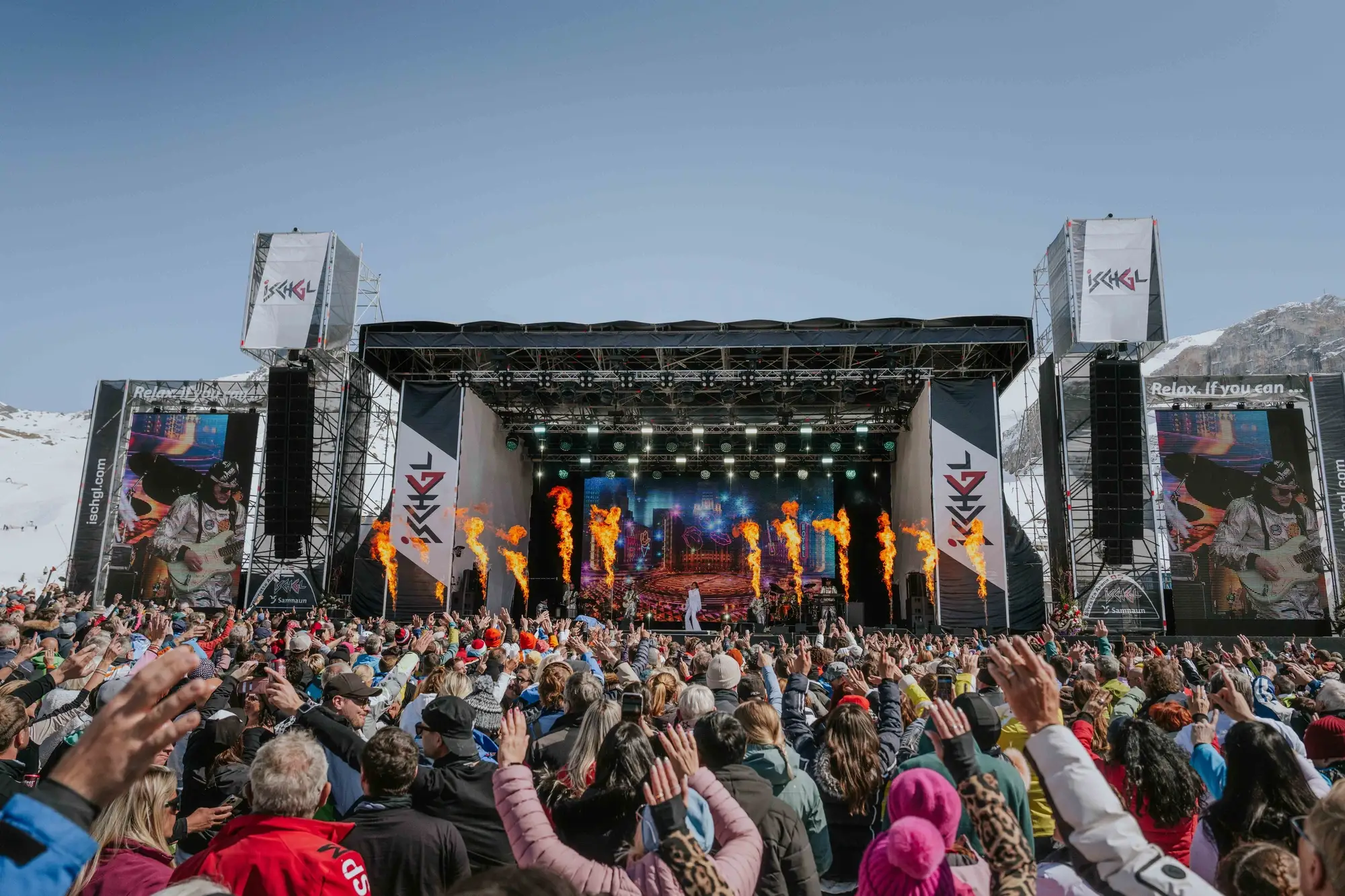 Konzert im Freien mit Publikum, Bühne, Flammen und Bergen im Hintergrund.