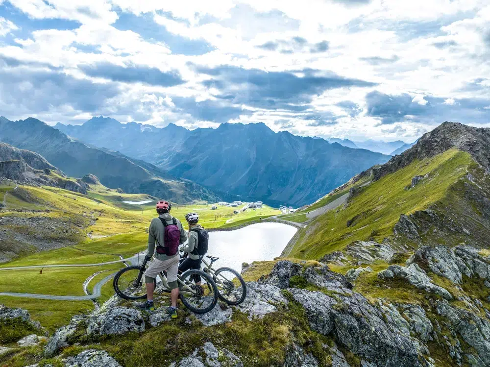 Zwei Radfahrer mit Fahrrädern bewundern eine malerische Berglandschaft mit einem See und verschlungenen Pfaden.