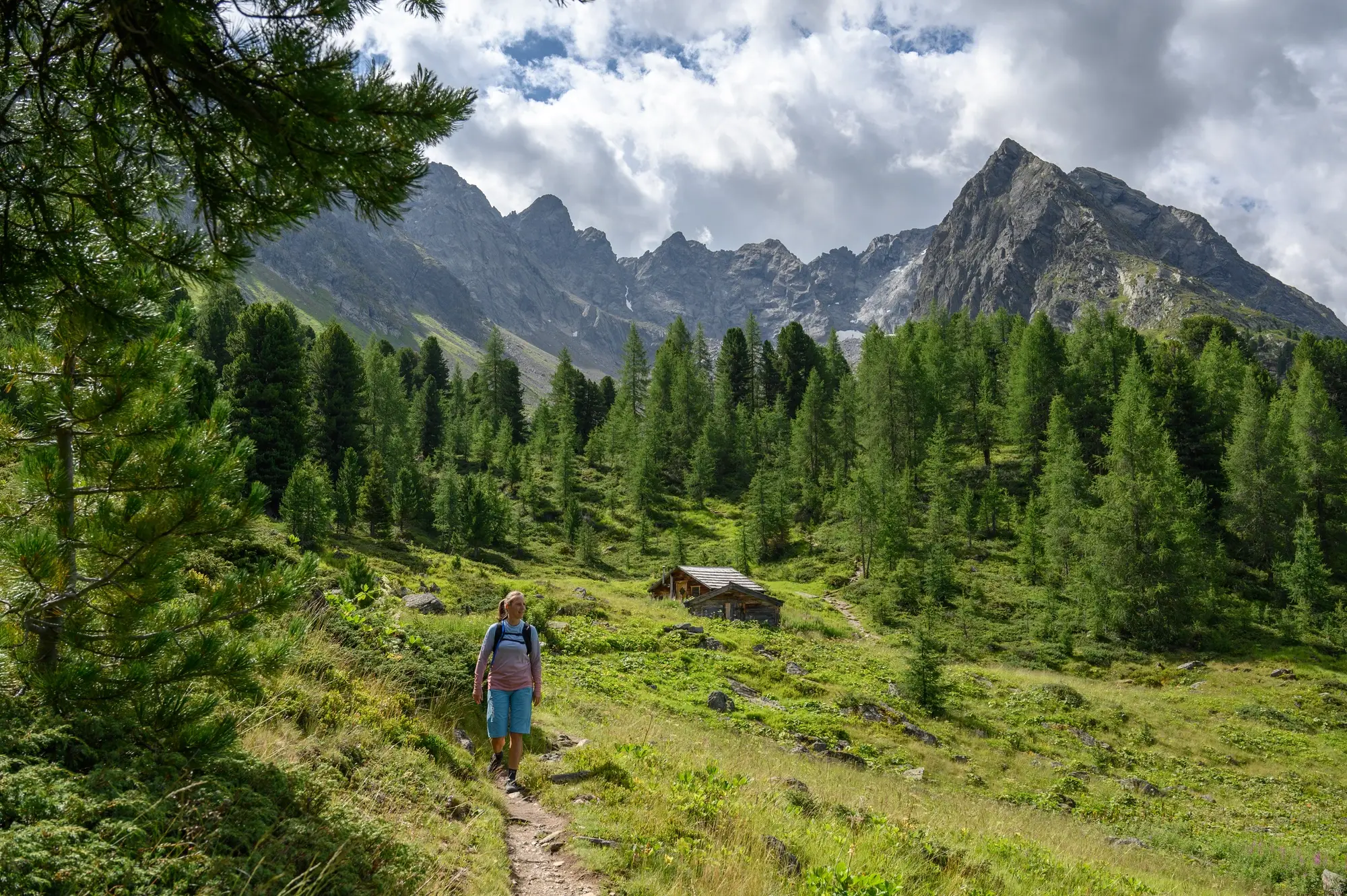 Person beim Wandern auf einem Bergpfad mit Kiefern, einer Hütte und schroffen Gipfeln im Hintergrund.