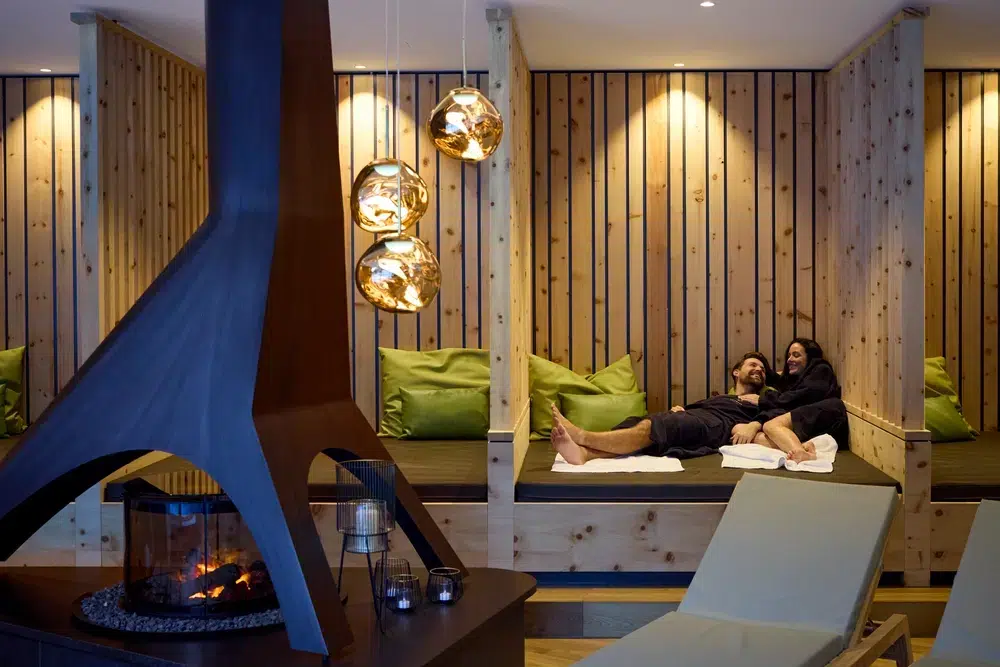 Ein Paar entspannt sich auf einem Lounge-Bett in einem gemütlichen, holzgetäfelten Zimmer mit modernem Kamin.