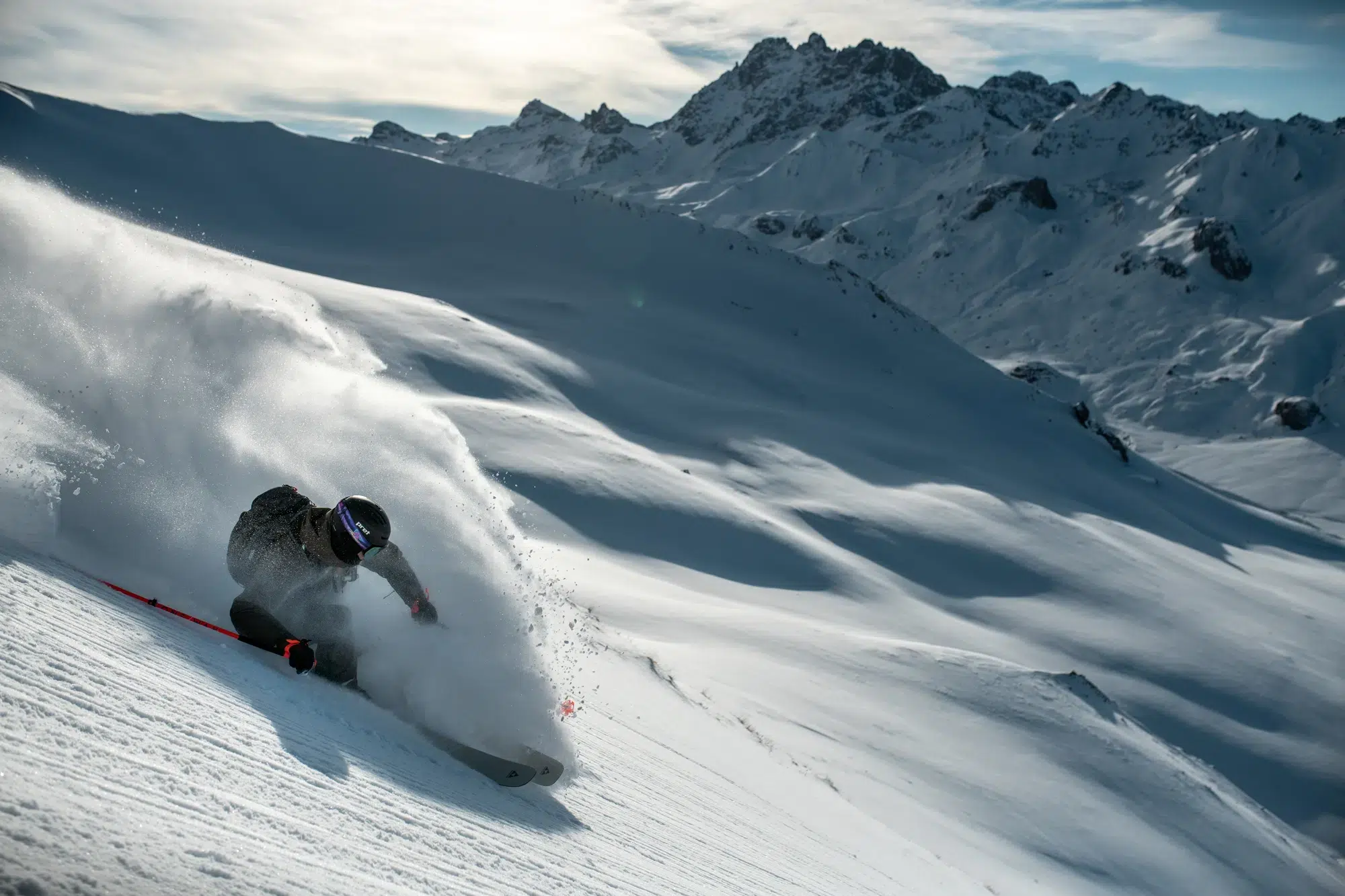Skifahrer, der auf einem verschneiten Berghang mit Gipfeln im Hintergrund durch den Pulverschnee carvt.
